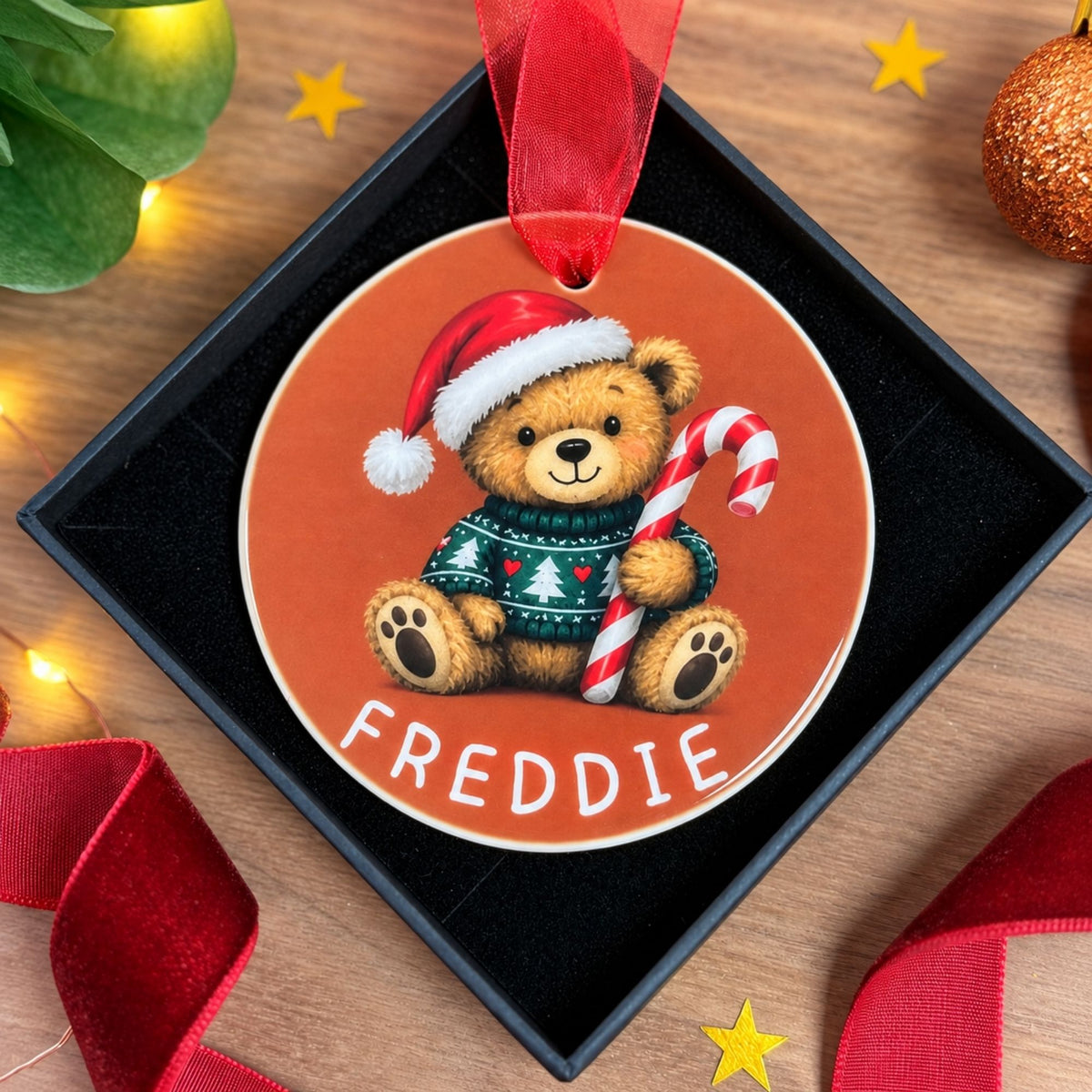 A round Christmas ornament featuring a teddy bear in a red Santa hat and green sweater holding a candy cane, with the name 'FREDDIE' printed at the bottom. The ornament hangs from a red ribbon inside a black gift box on a wooden surface with holiday decorations around it.