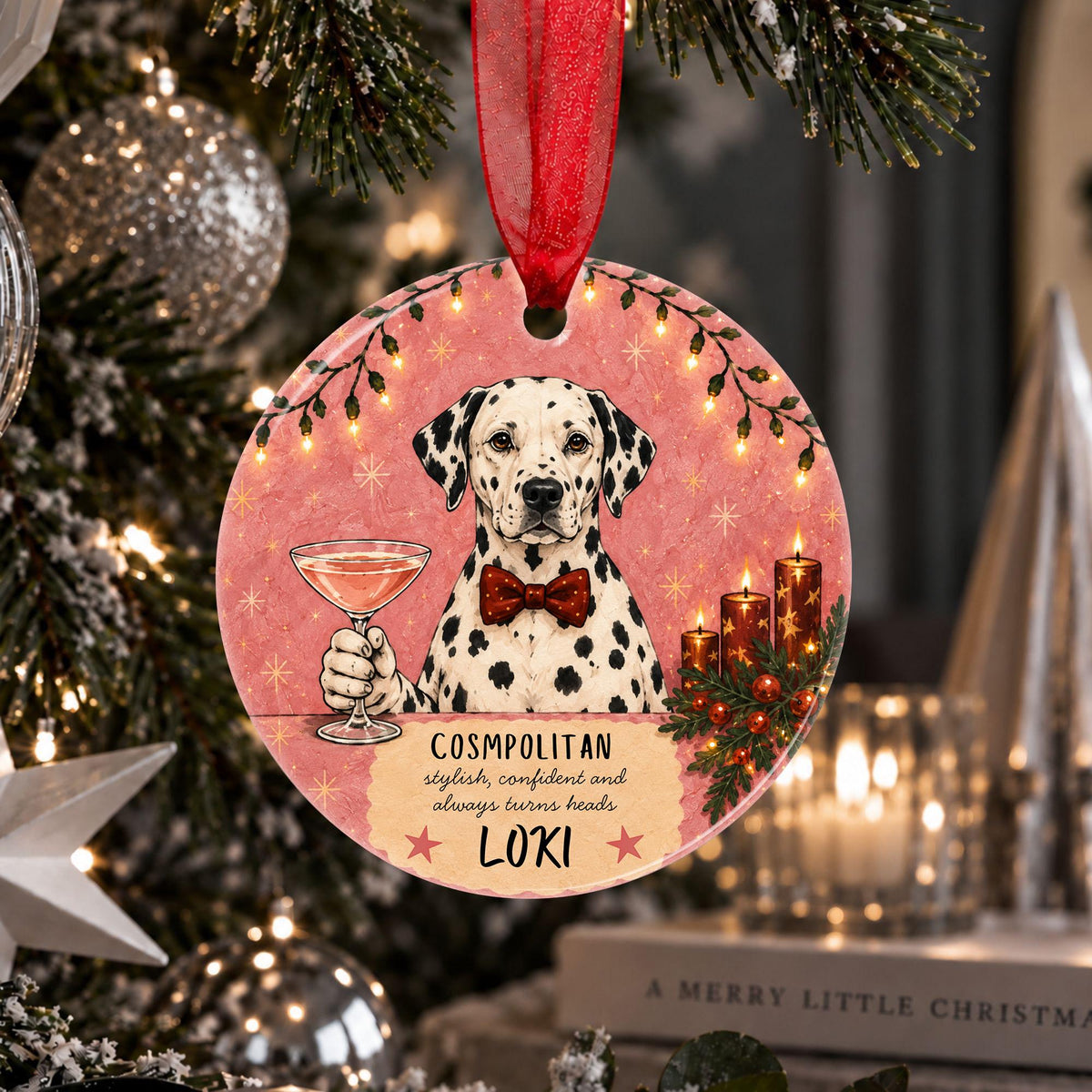 A round Christmas ornament hanging from a red ribbon on a decorated Christmas tree. The ornament features an illustration of a Dalmatian wearing a red bow tie, holding a cocktail glass, with pink background, holiday lights, candles, and the text 'COSMOPOLITAN stylish, confident and always loves dogs LOKI'.