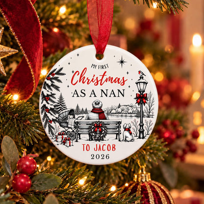 A personalized round Christmas ornament hanging on a decorated tree. The white ornament features black line art of a holiday scene with a bench, dog, lamp post, and red ribbon accents, along with the text 'MY FIRST Christmas AS A NAN TO JACOB 2026'.