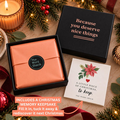 A Christmas-themed gift set from The Word Snack, featuring a black gift box with rose gold lettering, a peach-colored wrapped keepsake, a white card with a red poinsettia, and a candle on a rustic holiday background.