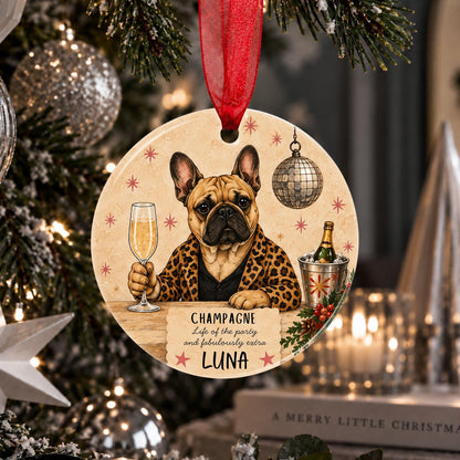 A personalized round Christmas ornament featuring a French Bulldog in a leopard-print shirt holding a glass of champagne, with the name 'LUNA' and the phrase 'Champagne Life of the party and fabulously extra' printed on it. The ornament hangs from a red ribbon in front of a decorated Christmas tree.