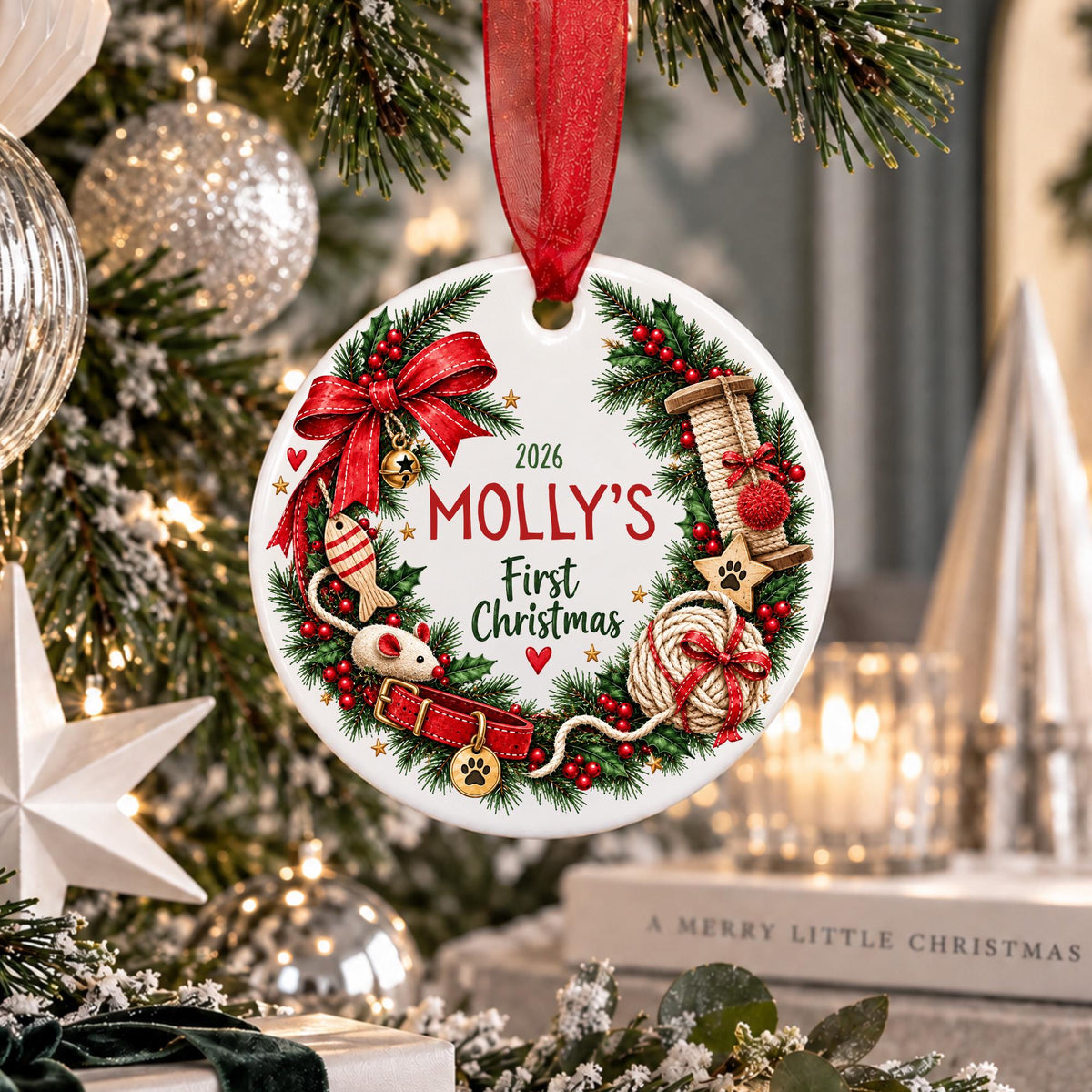 A round white Christmas ornament hanging from a red ribbon, decorated with a green wreath, red bow, red text reading '2026 MOLLY'S First Christmas,' and holiday illustrations including a red gift box, paw prints, and a spool of thread. The ornament is photographed in a festive indoor setting with a lit Christmas tree and holiday decor in the background.