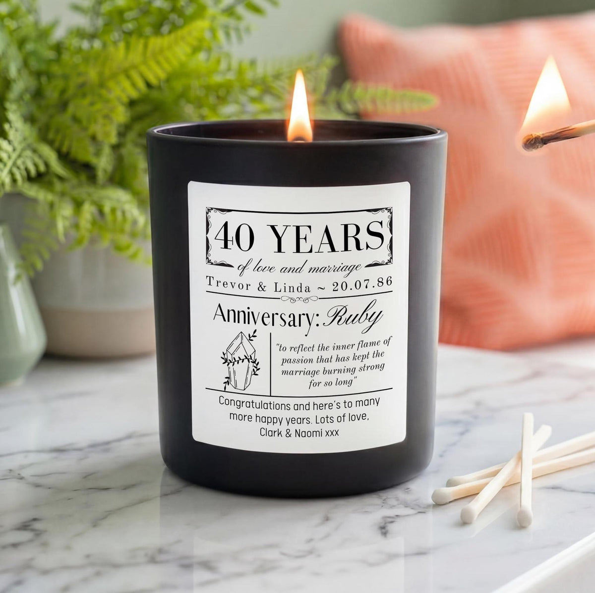 A black ceramic candle jar with a white printed anniversary label reading '30 Years of love and marriage' and 'Anniversary: Pearl,' shown lit on a marble surface with matches, a plant, and a pink cushion in the background.