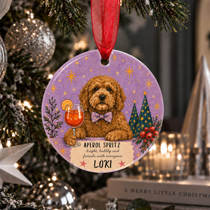 A round Christmas ornament hanging from a red ribbon in front of a decorated Christmas tree. The ornament features a cartoon illustration of a golden cockapoo wearing a purple bow tie, with a wine glass, pine trees, and the text 'APEROL SPRIT bright, bubbly and friends with everyone LOKI'.