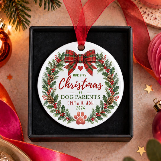 A personalized round Christmas ornament displayed inside a black gift box, featuring a white background with a red ribbon bow, green holly wreath, pinecones, and the text 'Our First Christmas as DOG PARENTS EMMA & JON 2026'. The box is styled on a warm beige surface with festive holiday decorations including pine branches, red ribbons, and ornaments.