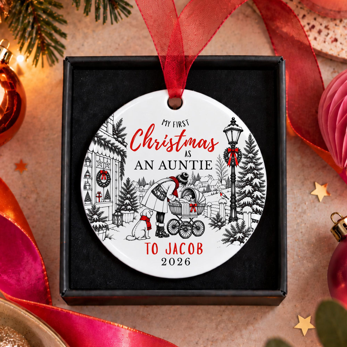 A personalized round Christmas ornament displayed in a black gift box, featuring a festive black and white illustration of a man pushing a stroller with a dog, red script text reading 'My First Christmas As An Auntie', and 'To Jacob 2026' at the bottom. The ornament hangs from a red ribbon, surrounded by holiday decorations like pine branches, ribbons, and ornaments.