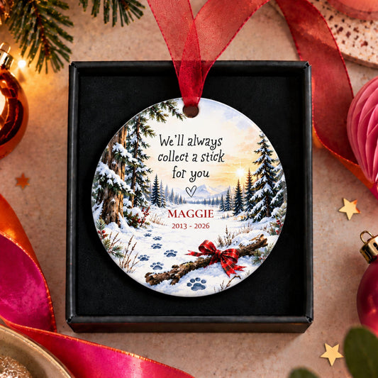 A personalized round Christmas ornament displayed inside a black gift box, surrounded by festive holiday decorations. The ornament features a winter forest scene with paw prints and the text 'We'll always collect a stick for you' and the name 'MAGGIE 2013 - 2026'.