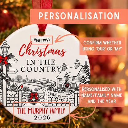 Personalized round Christmas ornament with a festive winter village illustration and text reading 'Our First Christmas in the Country' and 'The Murphy Family 2026', shown with promotional callouts on a warm blurred holiday background.