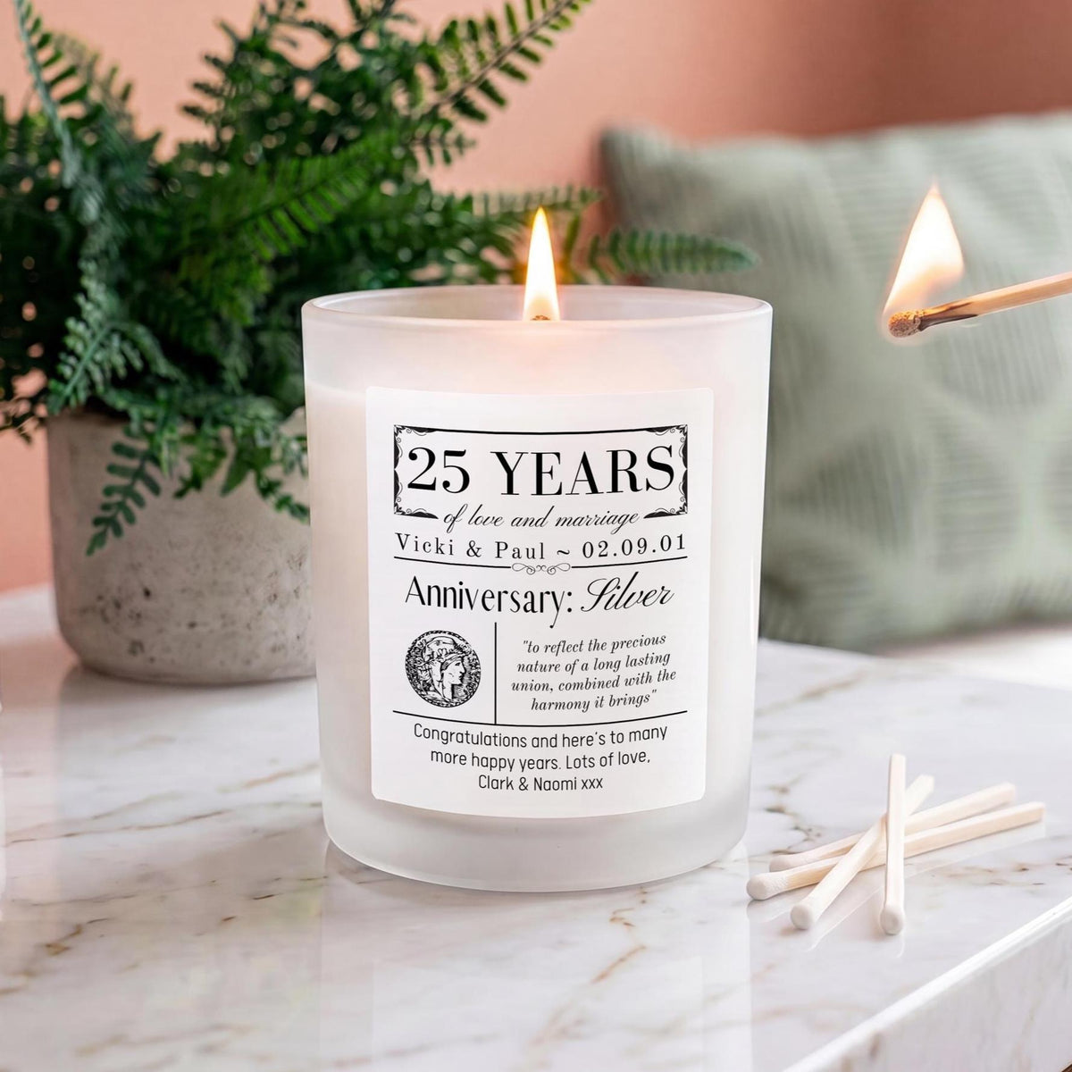 A frosted glass candle with a lit flame sits on a marble surface in a styled home setting, with a matchbook-style personalized label reading '25 Years Anniversary: Silver' and a matchbook with lit matches nearby, plus a plant and sofa in the background.