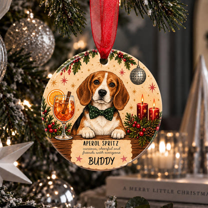 A round wooden Christmas ornament featuring a beagle dog wearing a green bow tie, hanging from a red ribbon in front of a decorated Christmas tree. The ornament includes festive illustrations of a drink, candles, pinecones, holly, and the personalized text 'APEROL SPRITZ sincere, cheerful and friendly with everyone BUDDY'.