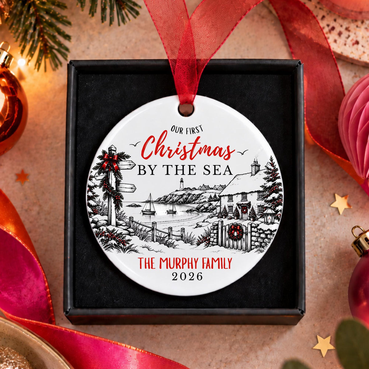 A personalized round Christmas ornament displayed in a black gift box, featuring a winter coastal illustration and the text 'Our First Christmas by the Sea' and 'The Murphy Family 2026'. The ornament has a red hanging ribbon and is surrounded by festive holiday decorations including ribbons, ornaments, and pine branches.