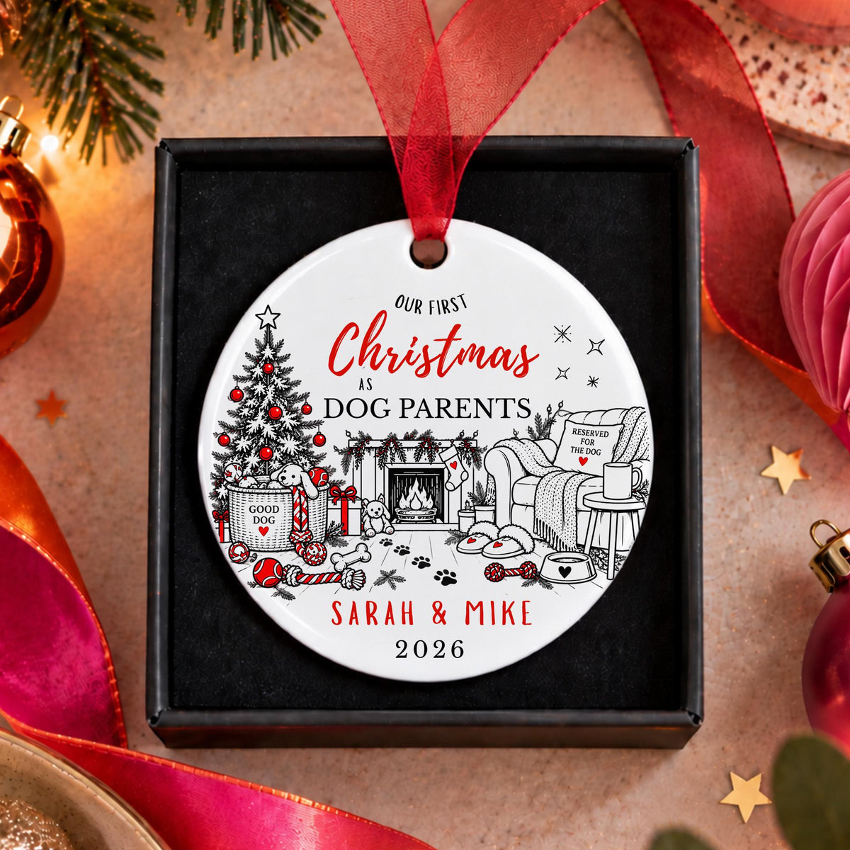 A personalized round Christmas ornament displayed in a black gift box, featuring a red ribbon and festive illustrated scene with the text 'Our First Christmas as Dog Parents Sarah & Mike 2026'.