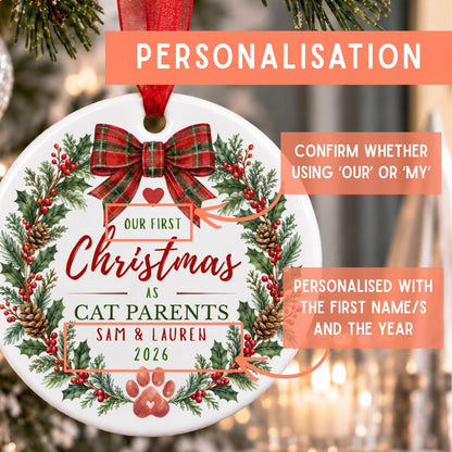 Personalized round Christmas ornament with a red ribbon hanger, featuring a festive wreath design with holly, pinecones, and red berries. The ornament includes customizable text such as 'Our First Christmas as Cat Parents' and sample names 'Sam & Lauren 2026'.