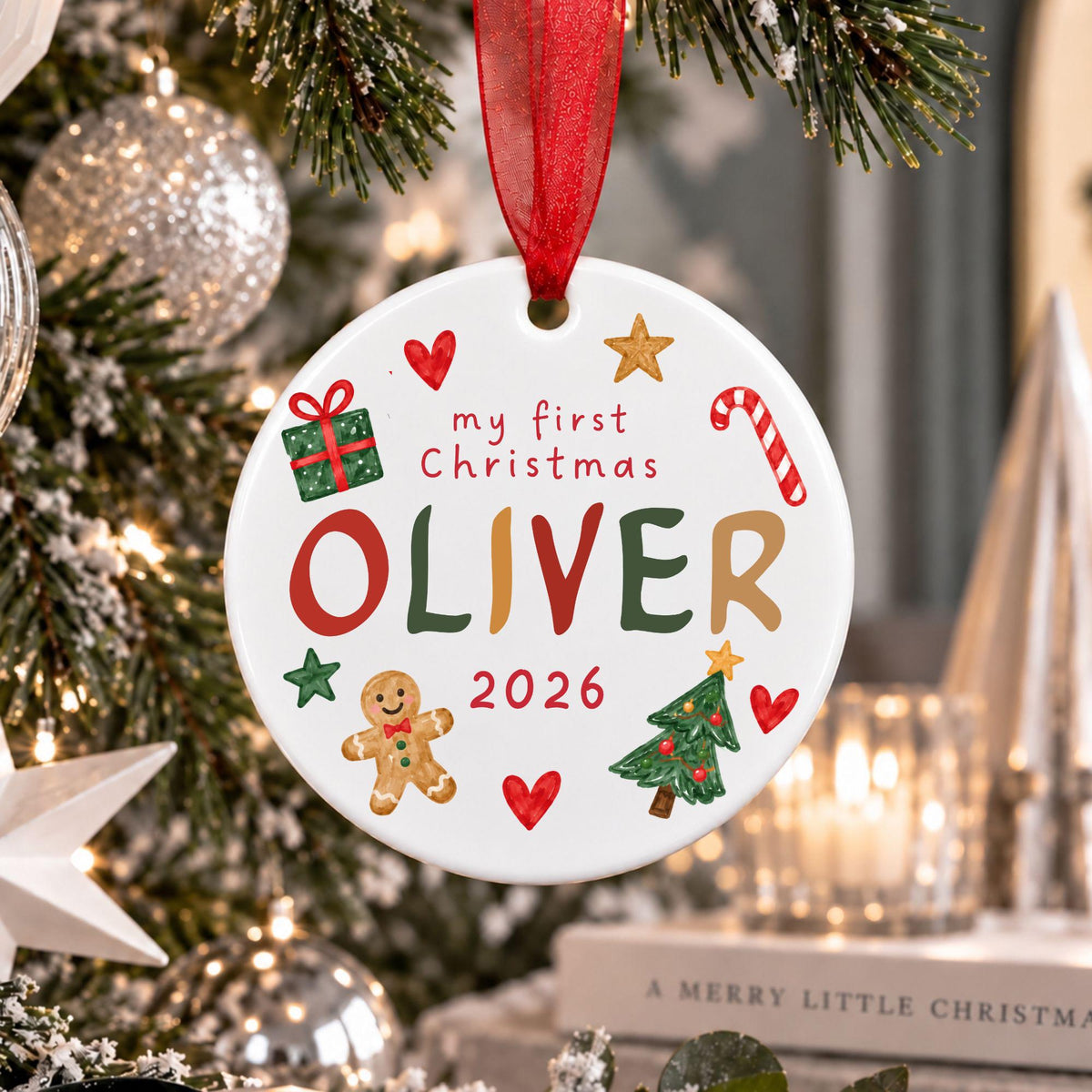 A personalized round white Christmas ornament hanging from a red ribbon on a decorated tree. The ornament features the text 'my first Christmas OLIVER 2026' with colorful illustrations of a gingerbread man, Christmas tree, candy cane, gift box, and hearts.