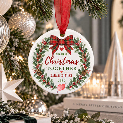 A personalized round ceramic Christmas ornament hanging from a red ribbon, featuring a festive wreath design with the text 'Our First Christmas Together Sarah & Mike 2026' on a decorated Christmas tree.