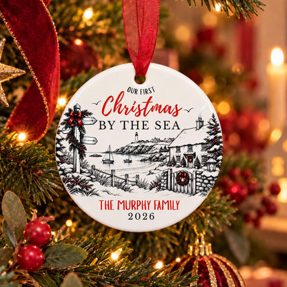 A round white ceramic Christmas ornament hanging on a decorated Christmas tree. The ornament features a black line art illustration of a coastal village and sea scene, with red and black text reading 'Our First Christmas by the Sea' and 'The Murphy Family 2026'.