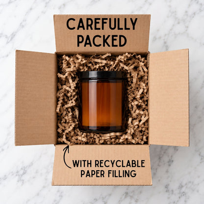 Amber glass scented candle with black lid securely packed inside a cardboard delivery box, surrounded by recyclable crinkle-cut paper filling for protection during shipping.