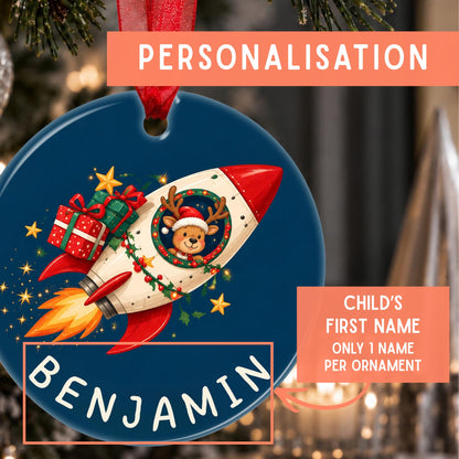 Personalized Christmas ornament featuring a blue round disc with a red ribbon loop, a white rocket carrying a Santa-clad reindeer, gift boxes, stars, and the name 'BENJAMIN' in white at the bottom. Coral text banners explain personalization and name customization.