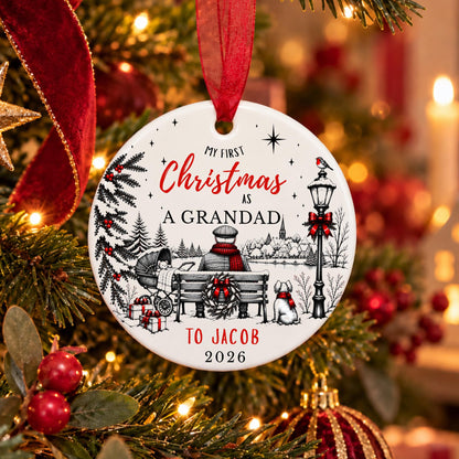 A round white Christmas ornament with a red hanging ribbon, featuring a festive black line drawing and the personalized text 'My First Christmas As A Grandad To Jacob 2026' in red and black. The ornament is hanging on a decorated Christmas tree with warm lights and red berries in the background.