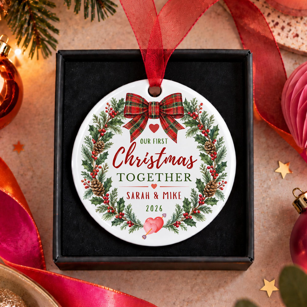 A personalized round Christmas ornament displayed inside a black gift box, surrounded by festive holiday decorations. The ornament features a white background with a red ribbon bow, a green pine wreath, and red and green text that reads 'Our First Christmas by the Sea' and 'The Murphy Family 2026'.