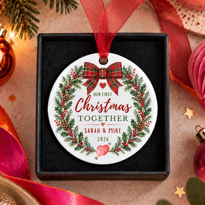 A personalized round Christmas ornament displayed inside a black gift box, surrounded by festive holiday decorations. The ornament features a white background with a red ribbon bow, a green pine wreath, and red and green text that reads 'Our First Christmas by the Sea' and 'The Murphy Family 2026'.