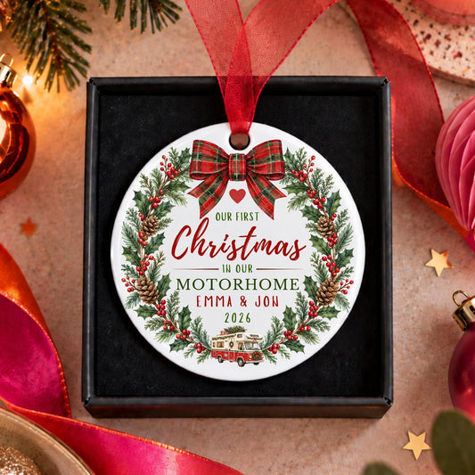 A personalized round Christmas ornament displayed in a black gift box, surrounded by festive holiday decorations. The ornament features a white background with a red ribbon loop, a red plaid bow, and a green holly wreath border. it says on it our first Christmas in our motorhome Emma & Jon 2026