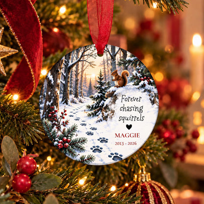 A personalized round Christmas ornament hanging on a decorated Christmas tree. The ornament features a snowy forest scene with paw prints, a squirrel on a pine tree, and the text 'Forever chasing squirrels' and 'MAGGIE 2013 - 2026'.