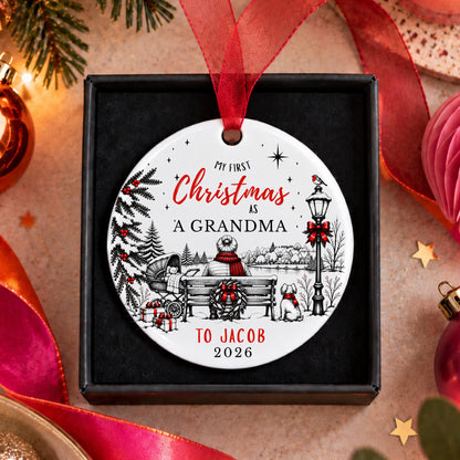 A personalized round Christmas ornament in a black gift box, featuring a winter scene with a bench, snowman, dog, and red ribbon text reading 'My First Christmas as a Grandma to Jacob 2026'. The ornament is surrounded by festive holiday decorations including pine branches, ribbons, and ornaments on a warm beige surface.