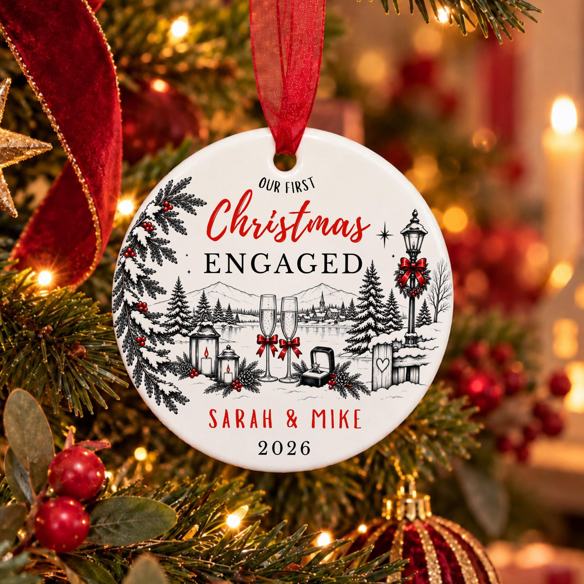 A round white Christmas ornament with a red ribbon hanger, featuring a black and red winter village illustration and the text 'Our First Christmas Engaged Sarah & Mike 2026,' hanging on a decorated Christmas tree with warm lights and red ornaments.