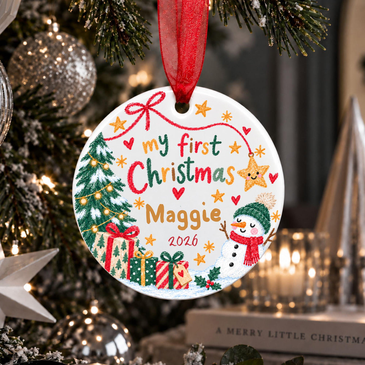 A personalized round Christmas ornament hanging from a red ribbon on a decorated Christmas tree. The white ornament features colorful hand-painted style illustrations and text reading 'my first Christmas Maggie 2026' with a tree, gifts, snowman, and stars.