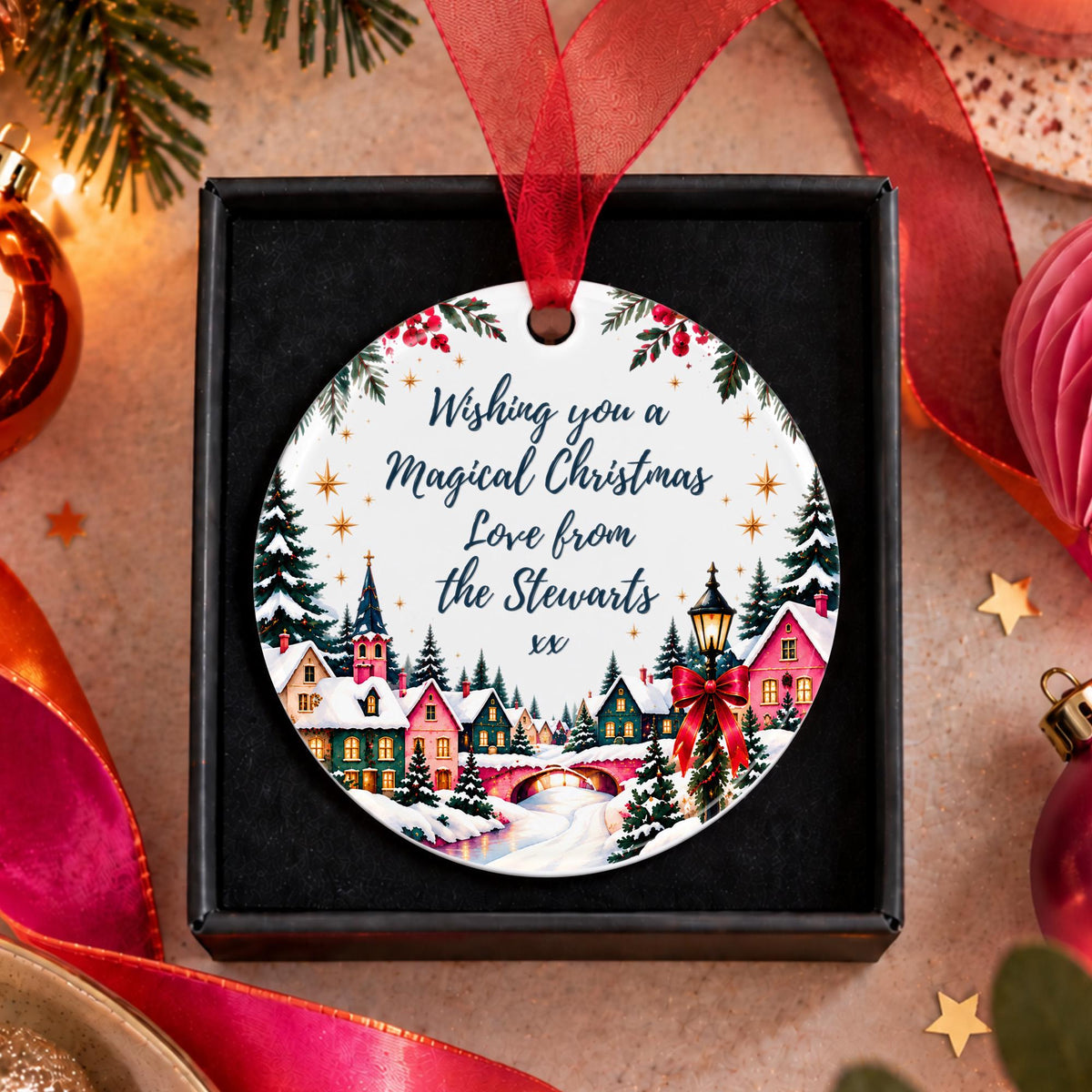 A round personalized Christmas ornament displayed in a black gift box, featuring a festive village illustration and the text 'Wishing you a Magical Christmas Love from the Stewarts xx'. The ornament hangs from a red ribbon, surrounded by holiday decorations like pine branches, ornaments, and ribbons on a warm beige background.