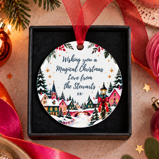 A round personalized Christmas ornament displayed in a black gift box, featuring a festive village illustration and the text 'Wishing you a Magical Christmas Love from the Stewarts xx'. The ornament hangs from a red ribbon, surrounded by holiday decorations like pine branches, ornaments, and ribbons on a warm beige background.
