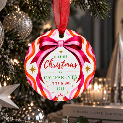 A personalized round Christmas ornament hanging on a decorated Christmas tree. The ornament features a colorful striped border in pink, orange, red, and white with a red bow graphic, and custom text reading 'Our First Christmas as Cat Parents Emma & Jon 2026'.