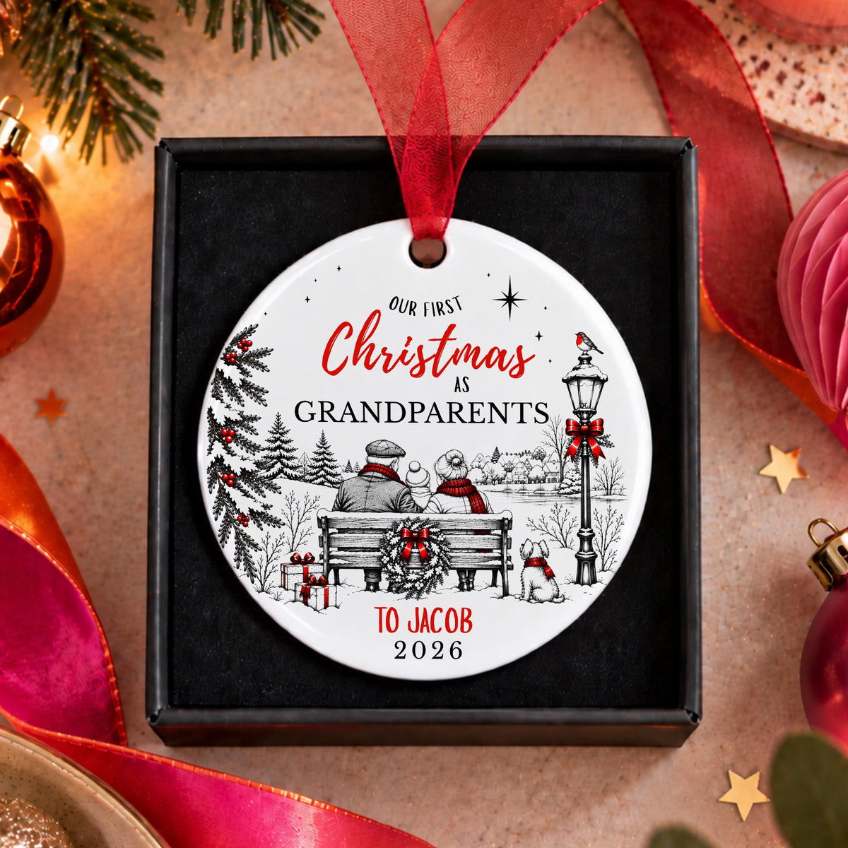 Personalized round Christmas ornament in a black gift box, featuring a winter holiday illustration with a couple on a bench, dogs, evergreen trees, and red ribbon text reading 'Our First Christmas as Grandparents to Jacob 2026'.