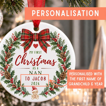 Personalized round Christmas ornament hanging on a tree, featuring a red plaid bow, green holly wreath, pinecones, and red berries. The ornament is printed with the text 'MY FIRST Christmas AS A NAN TO JACOB 2026' and includes promotional text overlays highlighting personalization.
