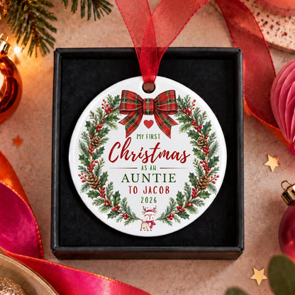 A personalized round Christmas ornament displayed in a black gift box, surrounded by festive holiday decorations. The ornament features a white background with a red ribbon bow, a green pine wreath, and red and green text that reads 'MY FIRST Christmas AS AN AUNTIE TO JACOB 2026'.