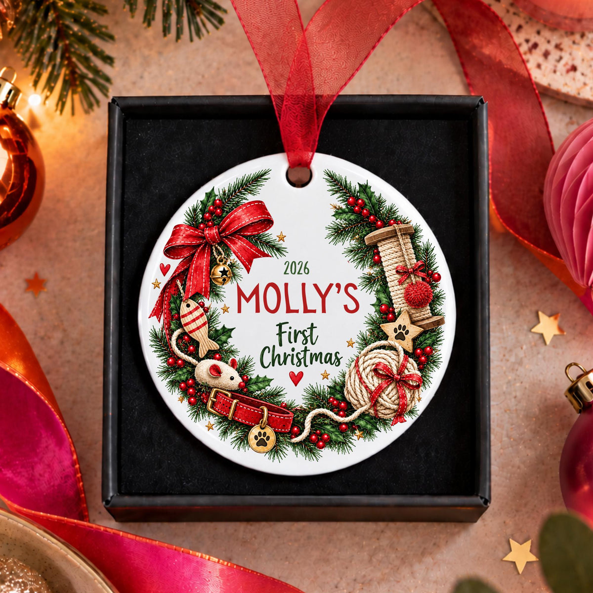 A personalized round Christmas ornament for pets, featuring a festive wreath design with red ribbon, green pine branches, and holiday illustrations. The ornament is printed with the text '2026 MOLLY'S First Christmas' and is displayed inside a black gift box with a red ribbon loop, surrounded by holiday decorations.