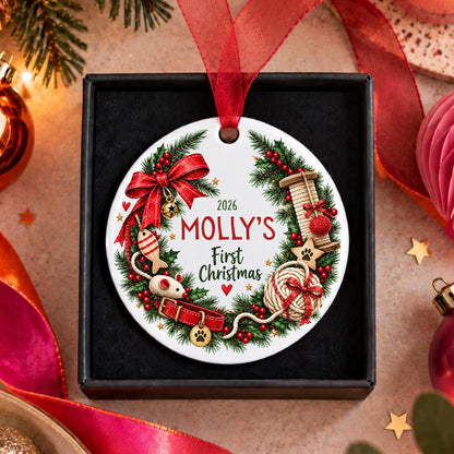 A personalized round Christmas ornament for pets, featuring a festive wreath design with red ribbon, green pine branches, and holiday illustrations. The ornament is printed with the text '2026 MOLLY'S First Christmas' and is displayed inside a black gift box with a red ribbon loop, surrounded by holiday decorations.