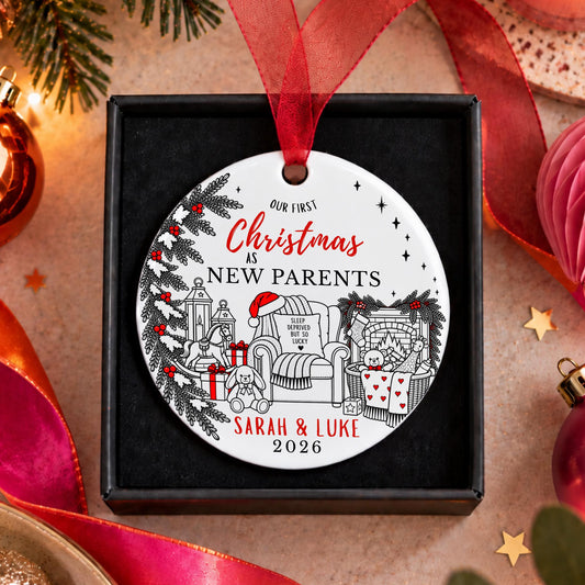 A personalized round Christmas ornament in a black gift box, featuring a white background with black line art of a cozy holiday scene and red text reading 'Our First Christmas as New Parents Sarah & Luke 2026'. The ornament hangs from a red ribbon, surrounded by festive holiday decorations including pine branches, ribbons, and ornaments.