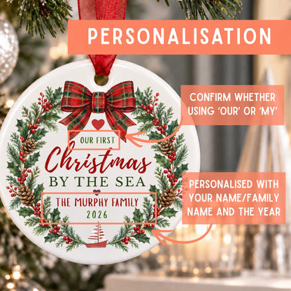 Personalized round Christmas ornament with a wreath design, red bow, and text reading 'Our First Christmas by the Sea' and 'The Murphy Family 2026,' shown hanging in front of a blurred Christmas tree with informational callouts about personalization.