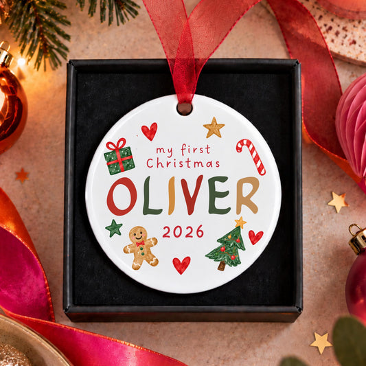 A personalized round ceramic Christmas ornament hanging from a red ribbon in a black gift box. The ornament is white with colorful festive illustrations and the text 'my first Christmas OLIVER 2026'.