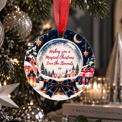 A personalized round Christmas ornament hanging from a red ribbon on a decorated Christmas tree. The ornament features a snow globe illustration with a winter village, gifts, and a blue ribbon bow, with the text 'Wishing you a Magical Christmas Love the Stewarts xx' in red script in the center.