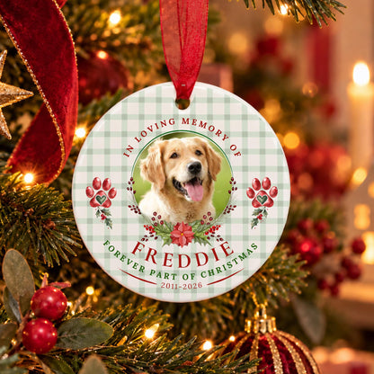 A personalized round Christmas ornament hanging from a red ribbon on a decorated Christmas tree. The ornament features a light green and white gingham pattern, red text reading 'IN LOVING MEMORY OF FREDDIE' and 'FOREVER PART OF CHRISTMAS 2011-2026', red paw prints, and a photo of a golden retriever dog in the center.