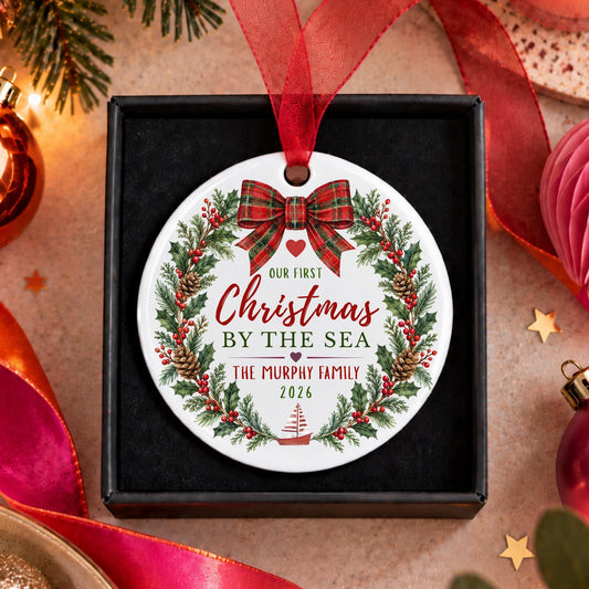 A personalized round Christmas ornament displayed inside a black gift box, surrounded by festive holiday decorations. The ornament features a white background with a red ribbon bow, a green pine wreath, and red and green text that reads 'Our First Christmas by the Sea' and 'The Murphy Family 2026'.