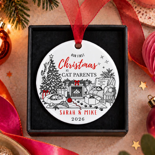 A personalized round Christmas ornament displayed in a black gift box, featuring a festive black line art illustration of a cat family scene with red text reading 'Our First Christmas as Cat Parents Sarah & Mike 2026'. The ornament hangs from a red ribbon, set against a warm holiday-themed background with ribbons, ornaments, and lights.