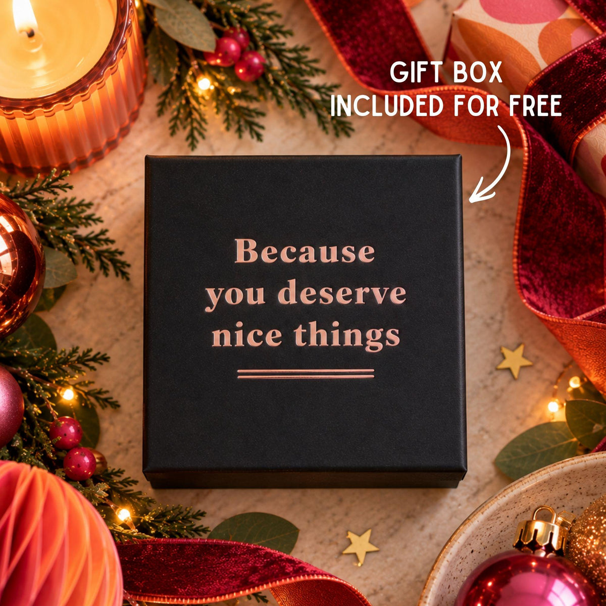 A square black gift box with rose-gold lettering that reads 'Because you deserve nice things,' surrounded by holiday decorations including candles, pine branches, ornaments, and red ribbon. White text in the top right says 'GIFT BOX INCLUDED FOR FREE' with an arrow pointing to the box.