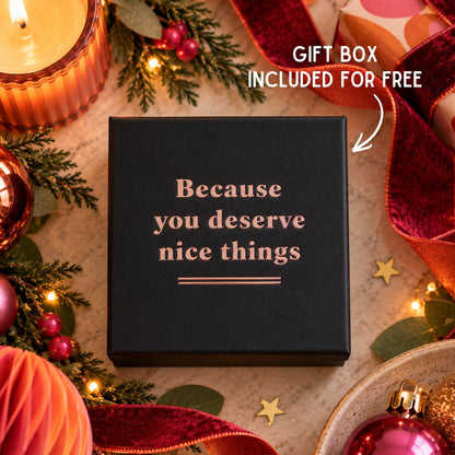 A square black gift box with rose-gold lettering that reads 'Because you deserve nice things,' surrounded by holiday decorations including candles, pine branches, ornaments, and red ribbon. White text in the top right says 'GIFT BOX INCLUDED FOR FREE' with an arrow pointing to the box.