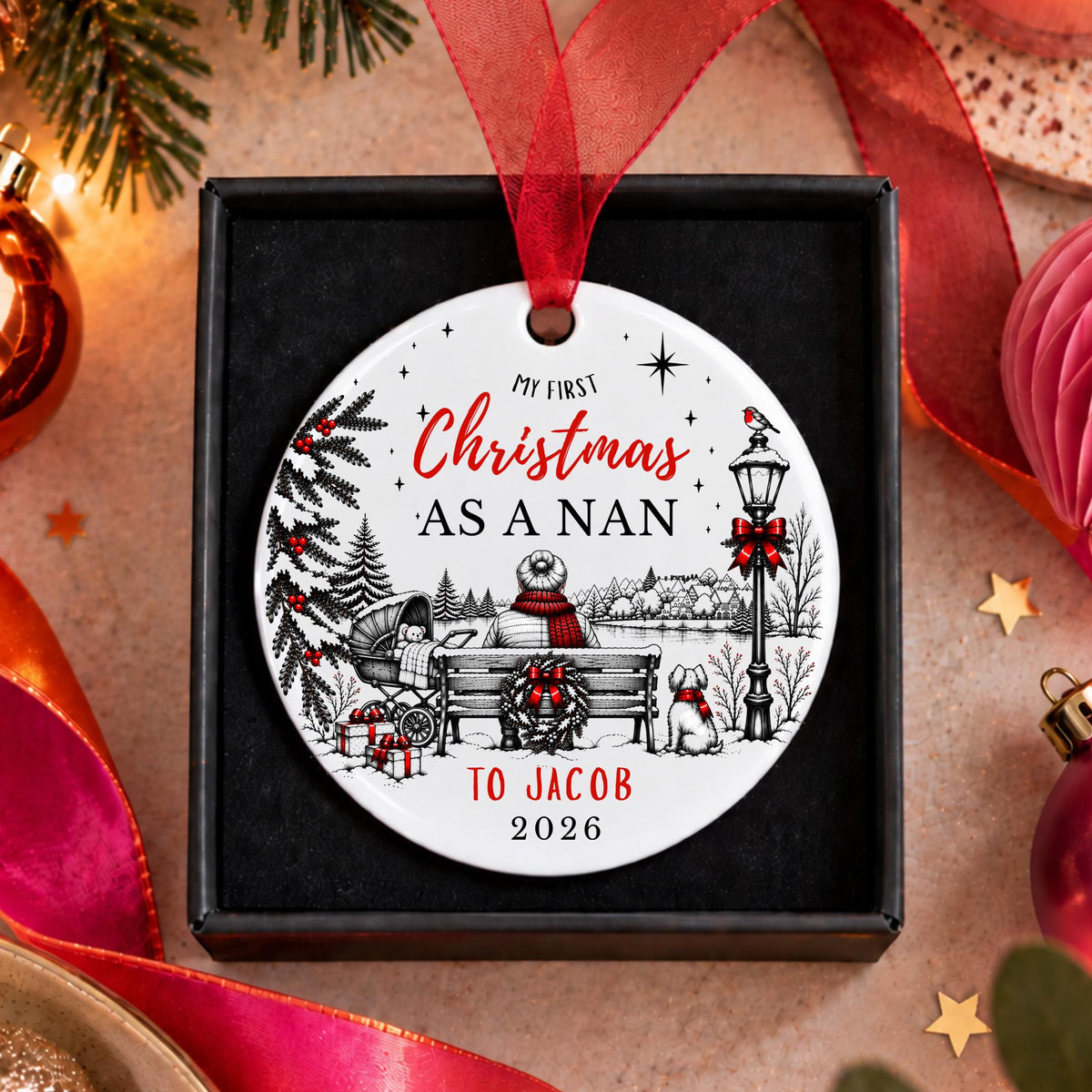A personalized round Christmas ornament displayed in a black gift box, featuring a red ribbon and festive winter illustration with the text 'MY FIRST Christmas AS A NAN TO JACOB 2026'.