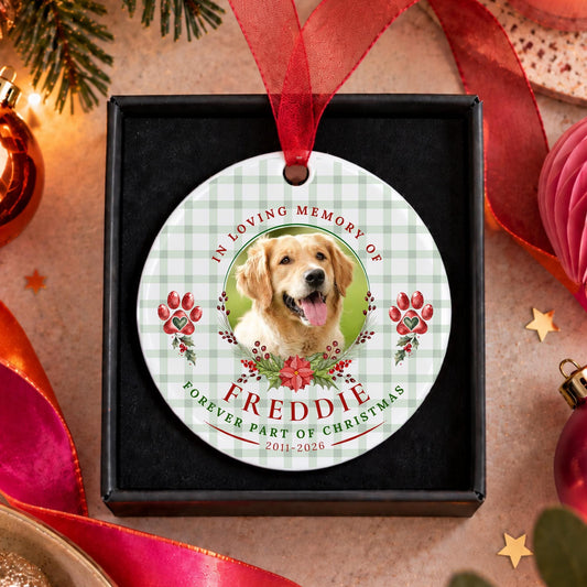 A personalized round Christmas ornament displayed in a black gift box, featuring a photo of a golden retriever dog with the text 'In Loving Memory of Freddie' and 'Forever Part of Christmas' on a white and green gingham background, surrounded by festive holiday decorations.