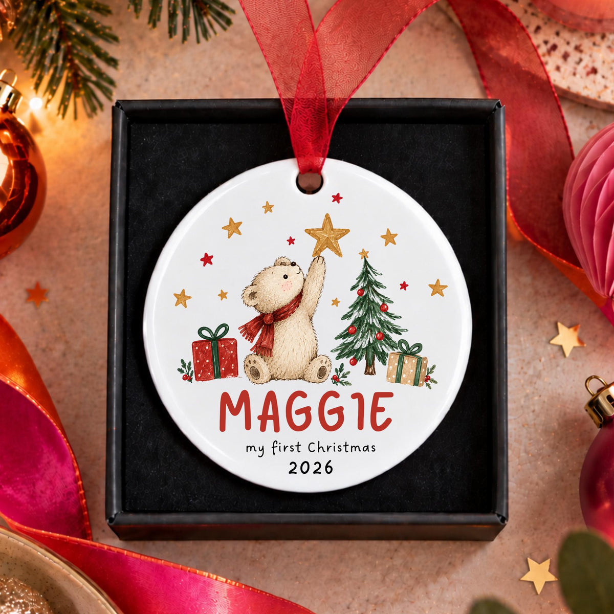 A personalized round Christmas ornament displayed in a black gift box, featuring an illustration of a teddy bear, a Christmas tree, gifts, and the name 'Maggie' with the text 'my first Christmas 2026'. The ornament has a red ribbon loop and is surrounded by festive holiday decorations.